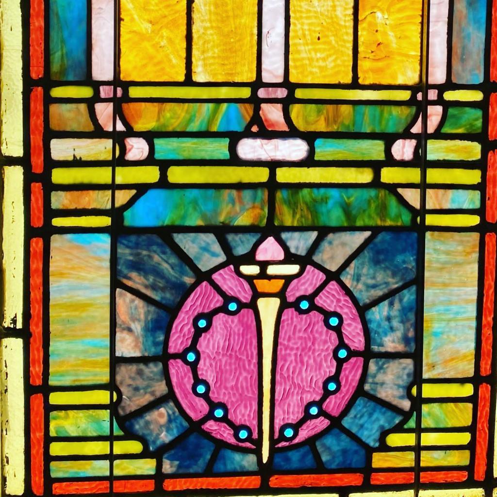 A portion of a stained glass window featuring a yellow, orange, and pink torch on top of a pink circle with blue dots. The window also features blue, green, and yellow blocks and a thin red rectangular border.